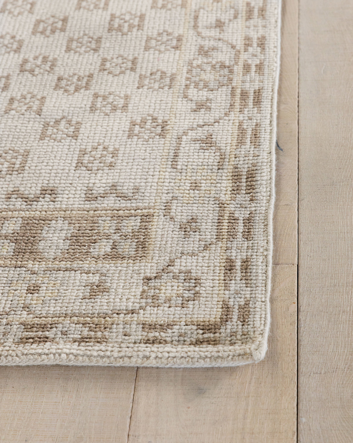 Close-up of the Anya Neutral Hand-Knotted Wool Rug in beige and light brown, featuring geometric and floral patterns. The detailed, durable woven texture is highlighted at the corner, displayed on a light wooden floor, by Vendixs