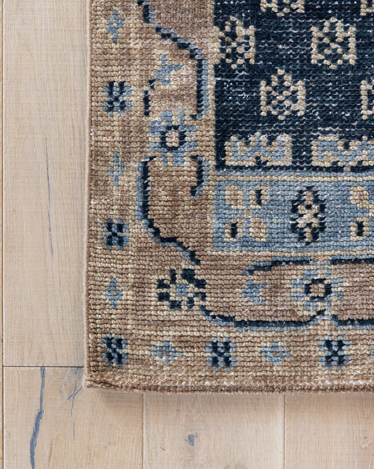 A close-up of the Anya Indigo Hand-Knotted Wool Rug featuring geometric and floral patterns in brown, black, and gray on a light wooden floor. Only the corner of this transitional wool rug is shown, by Vendixs