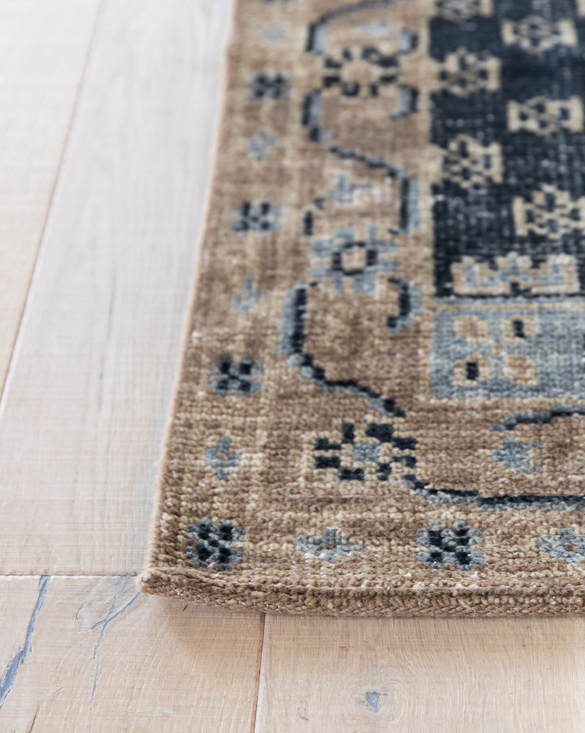 A close-up of the Anya Indigo Hand-Knotted Wool Rug shows its beige, black, and blue tones, textured weave, and transitional geometric border designs on a light wood floor, by Vendixs