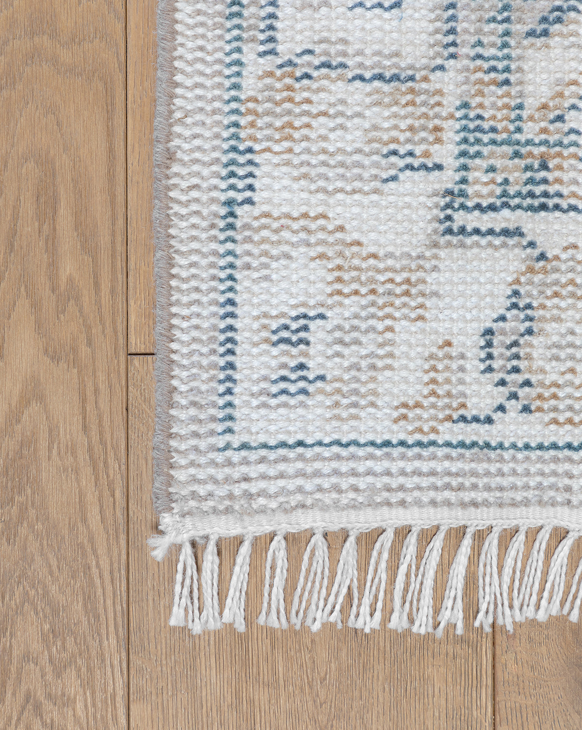 A close-up of the Merilyn Hand-Knotted Rug with white fringe, showcasing its subtle beige and blue pattern, displayed on a wooden floor, by Vendixs
