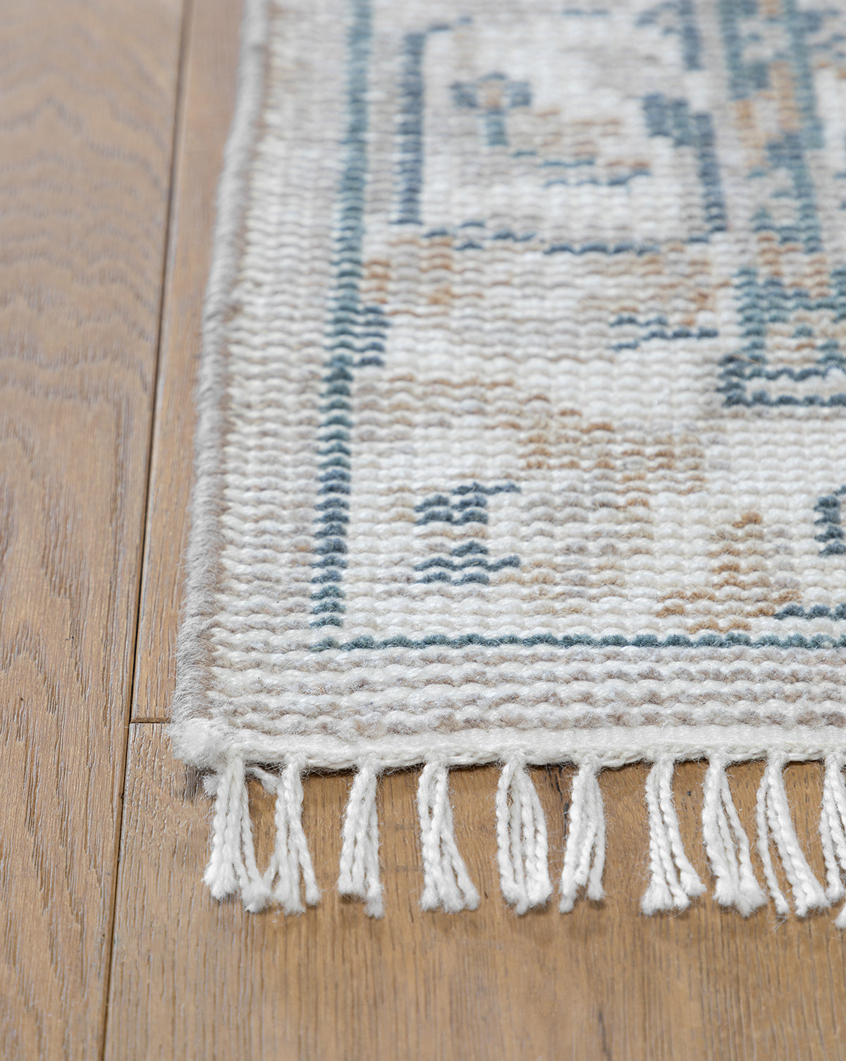 Close-up of the Merilyn Hand-Knotted Rug, featuring a beige and blue geometric pattern, white fringe accents, and fine craftsmanship. The rug is displayed elegantly on a light brown wooden floor, by McGee & Co.