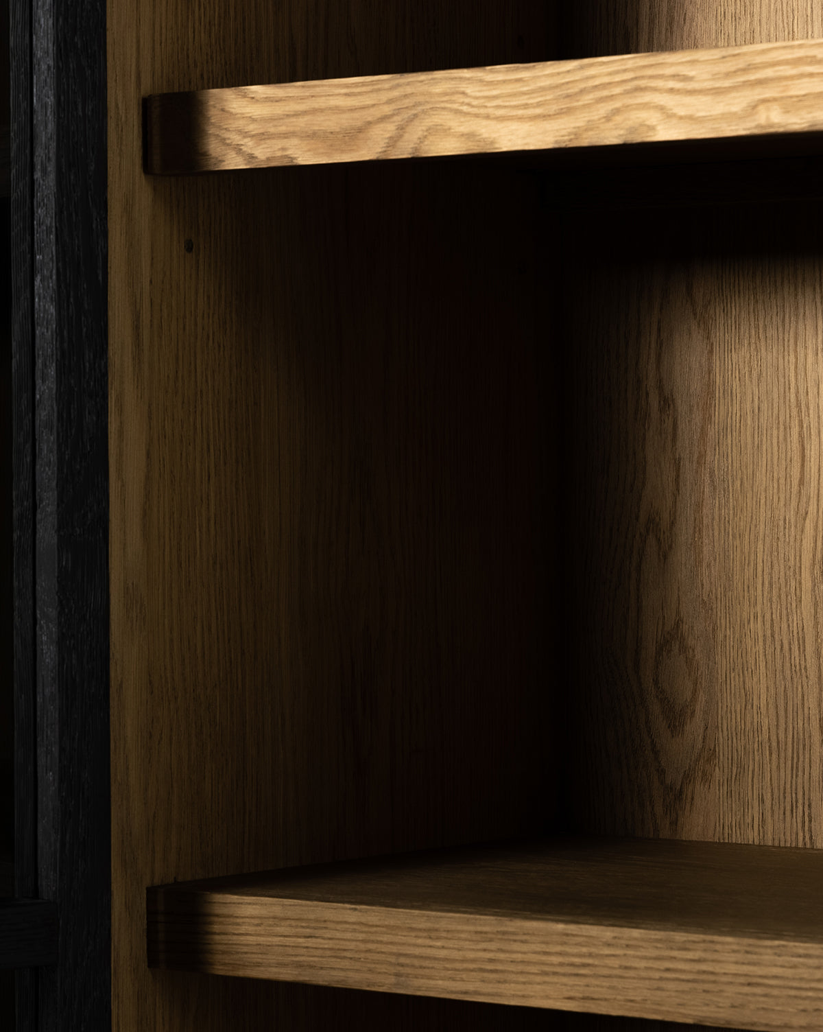 A close-up of three empty wooden shelves inside the Maddox Bookcase, showcasing natural wood grain and warm light highlighting the texture behind its elegant glass doors, by Vendixs