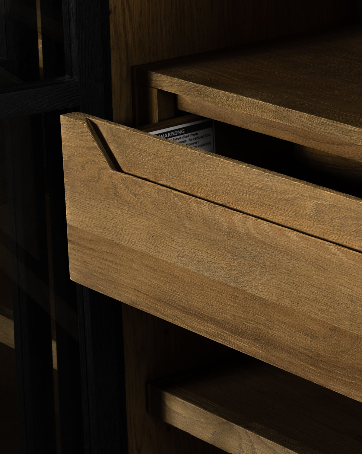 A partially opened drawer of the Maddox Bookcase reveals a warning label inside. The oak shelves and drawers are light brown, and a section of the black frame is visible on the left, by Vendixs