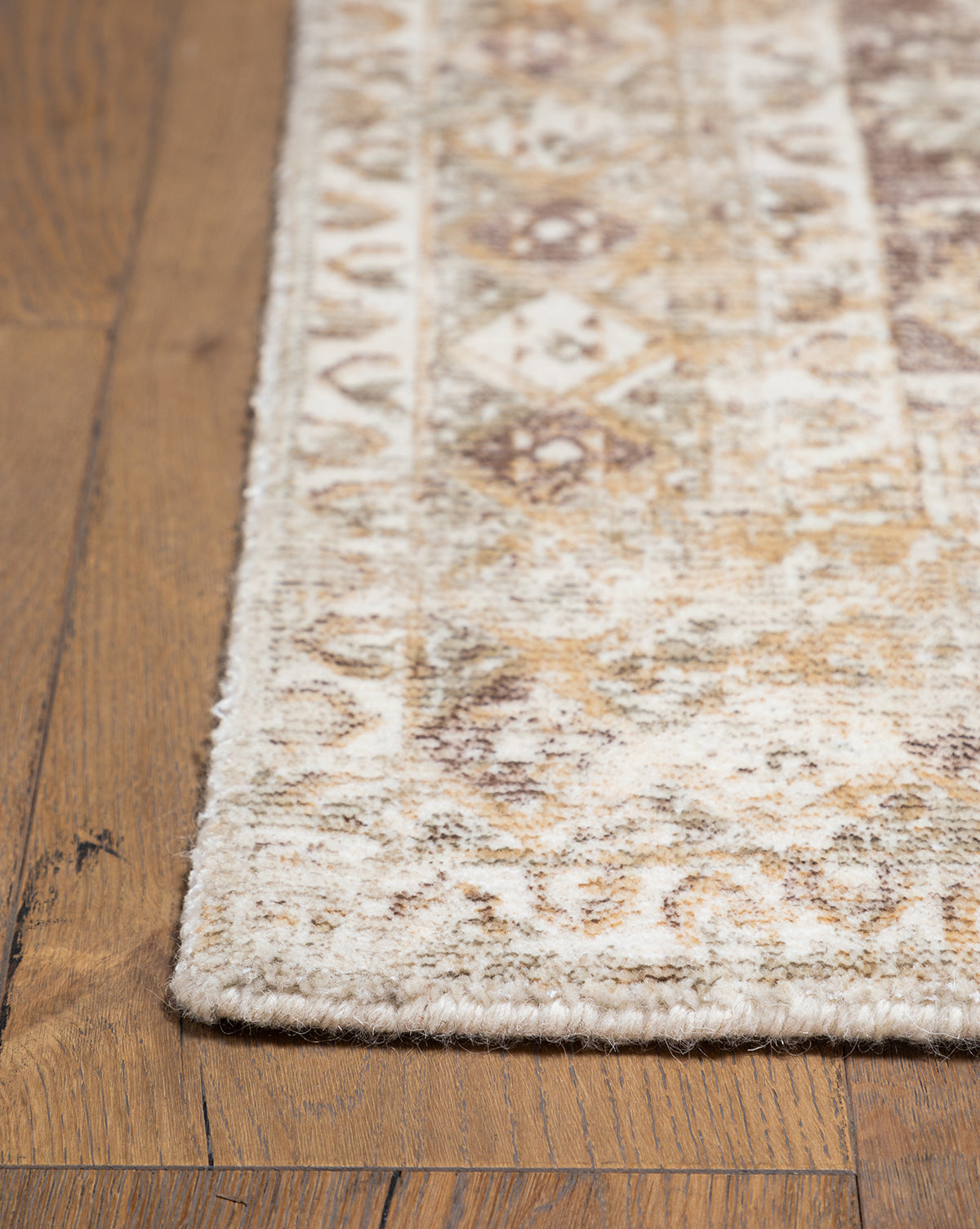 A close-up of the Marcella Handwoven Rug in neutral tan, beige, brown, and cream tones shows its subtle, faded geometric and floral pattern resting on a wooden floor, by Vendixs