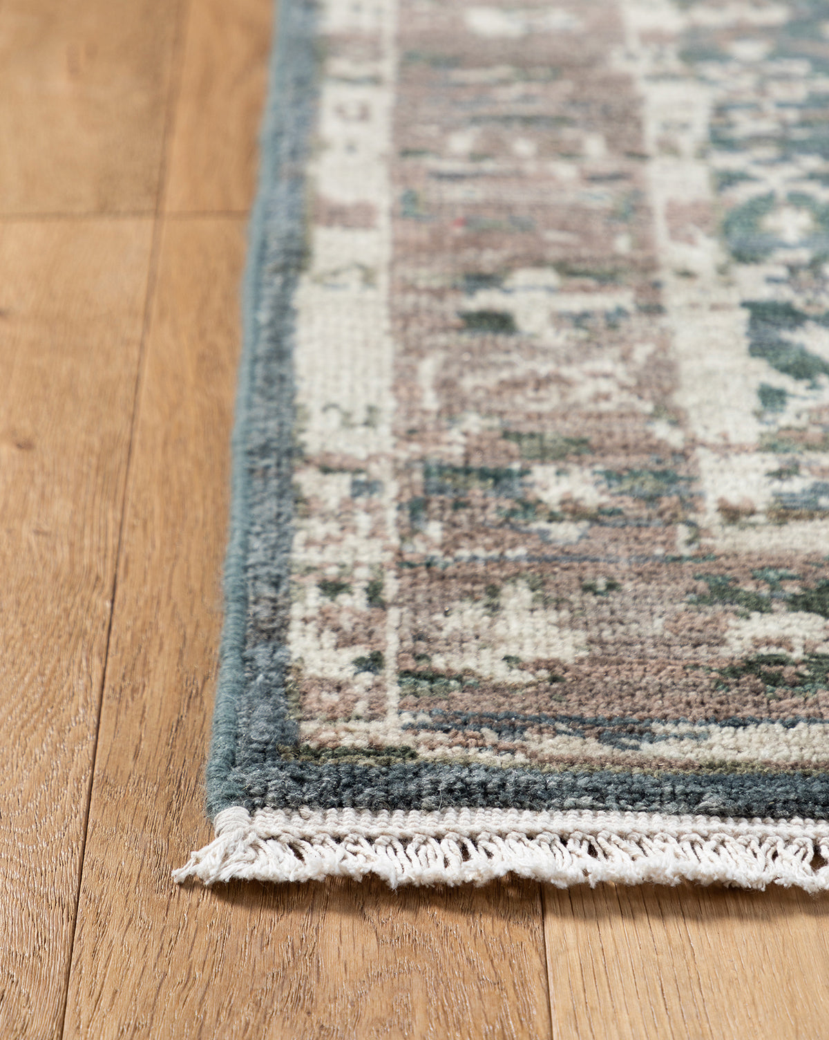 A close-up of the corner of the Marta Hand-Knotted Wool Rug, featuring blue, beige, and brown patterns with short white fringe, shown on a light wooden floor, by Vendixs