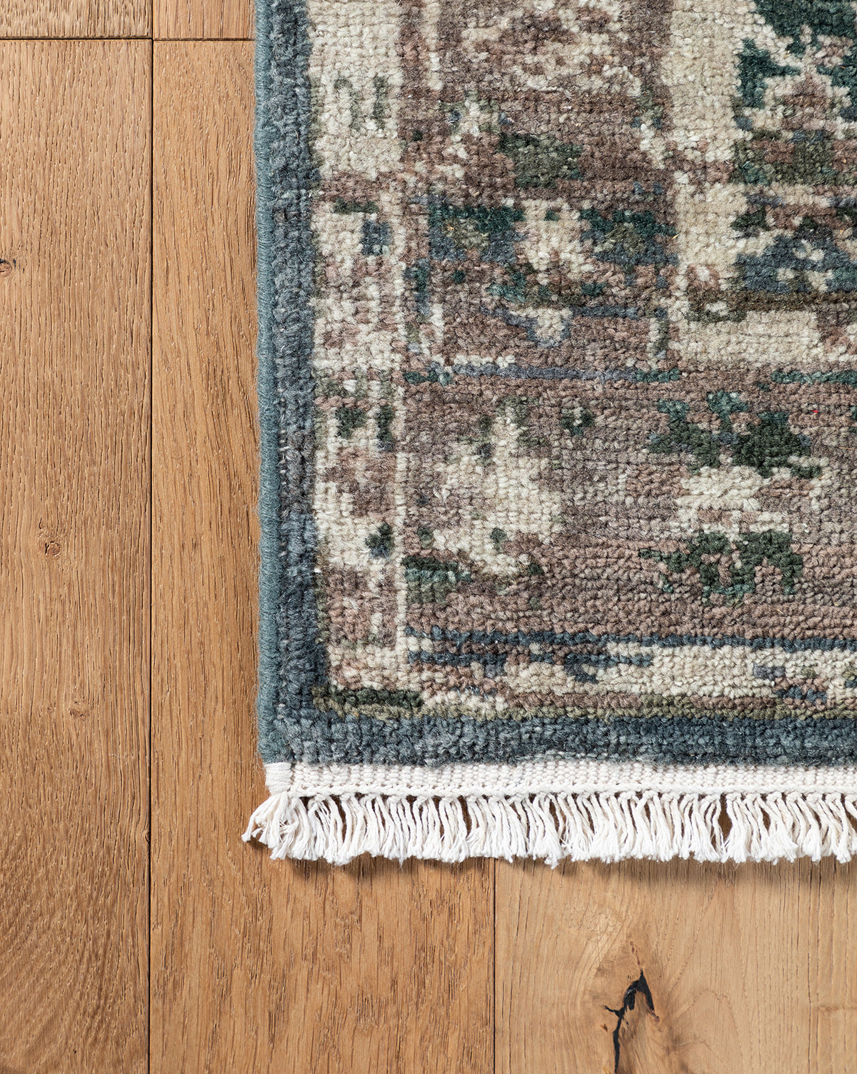 A close-up of the Marta Hand-Knotted Wool Rug with fringed edges on a wooden floor, showcasing its muted green, beige, and brown tones and worn vintage look, by Vendixs