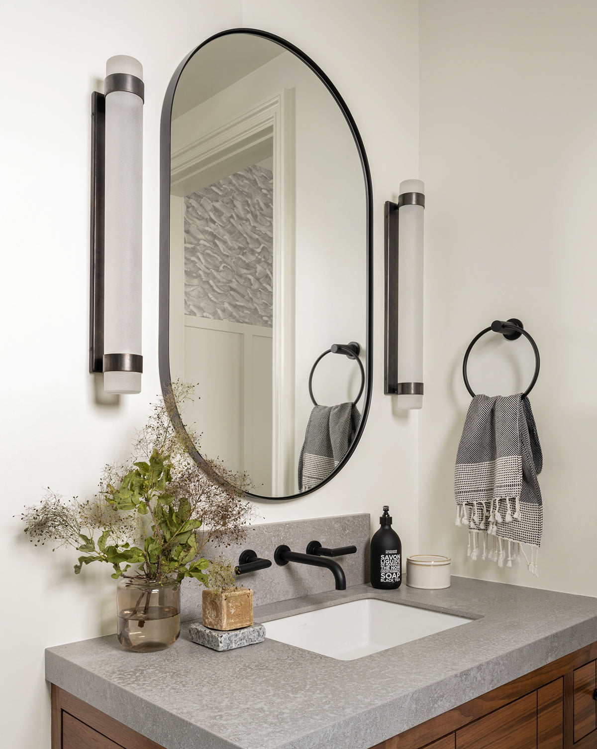 Modern bathroom vanity with the Rye Wall Mirror, vertical wall lights, gray countertop, rectangular sink, stainless steel soap dispenser, black faucet, potted plant, towel ring with gray towels, and decorative accents, by Vendixs