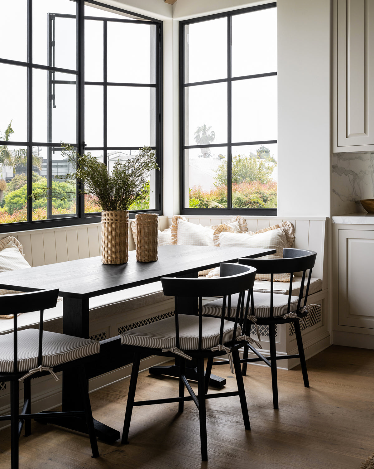 A modern dining area with a black table, Reeves Dining Chair, a built-in bench with cushions, large windows for natural light, and decorative vases filled with greenery on the table, by Vendixs
