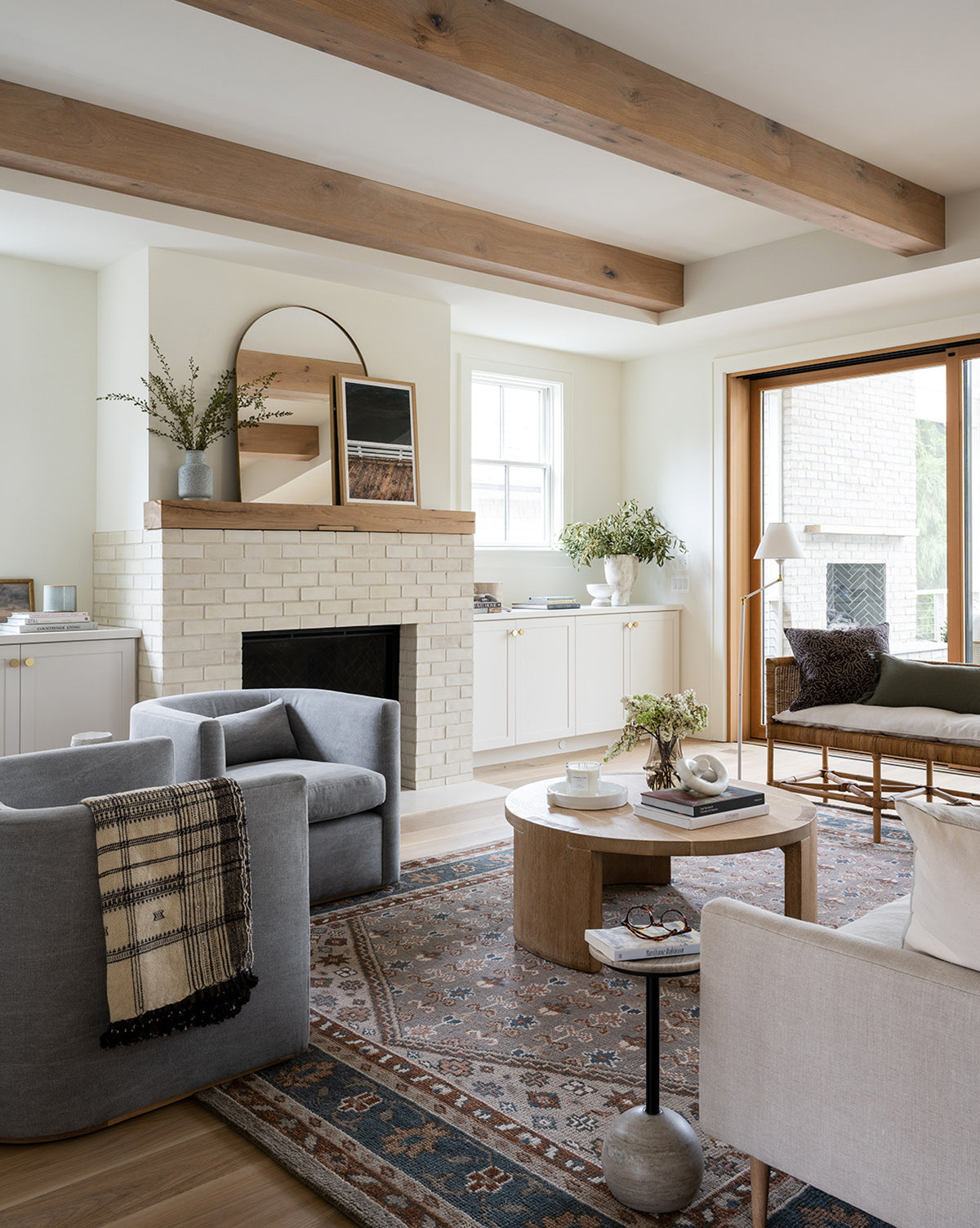 Bright, modern living room with wooden ceiling beams, white brick fireplace, neutral furniture, patterned rug, and the Raelynn Side Table with an antique marble top. Features coffee table, greenery, large windows, art, books, and a throw blanket by McGee & Co.