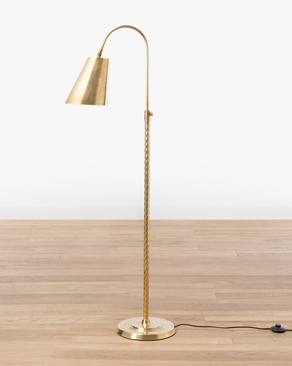 The Saylor Floor Lamp by Makerspalm features an elegant curved arm, conical shade, twisted brass stem, round base, and black power cord with switch—perfect for adding stylish lighting to your living room. Vendixs