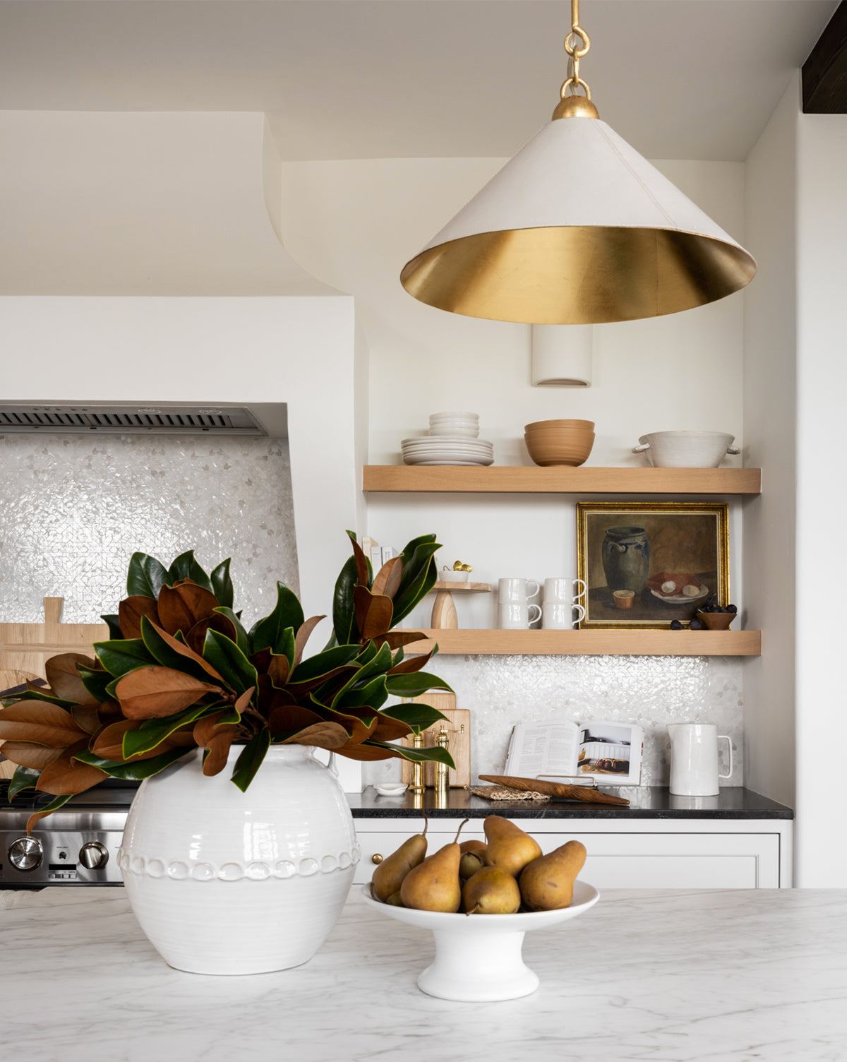 A modern kitchen features open wooden shelves with dishes and art, a white vase of magnolia leaves, a bowl of pears on marble, and elegant overhead lighting from the Scarlett Pendant, by Vendixs