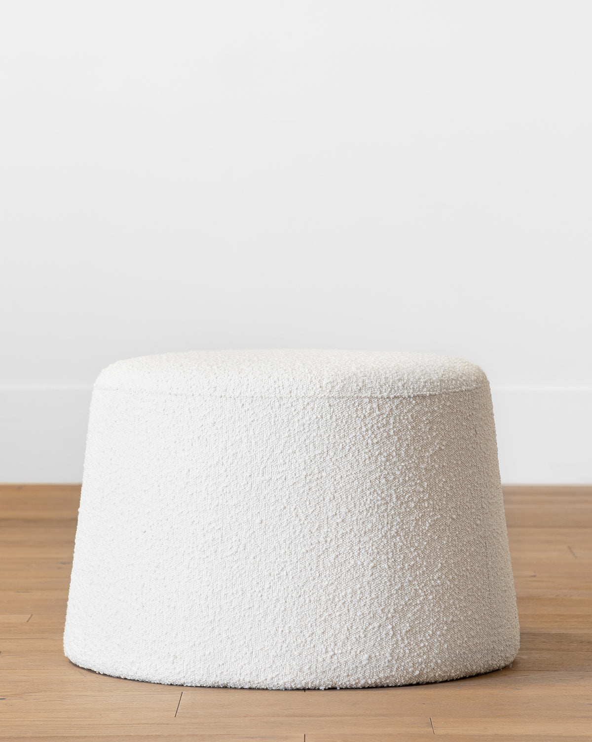 The Makerspalm Scout Ottoman features round, cream boucle upholstery with textured finish, resting on a light birch wood floor against a plain white wall by Vendixs
