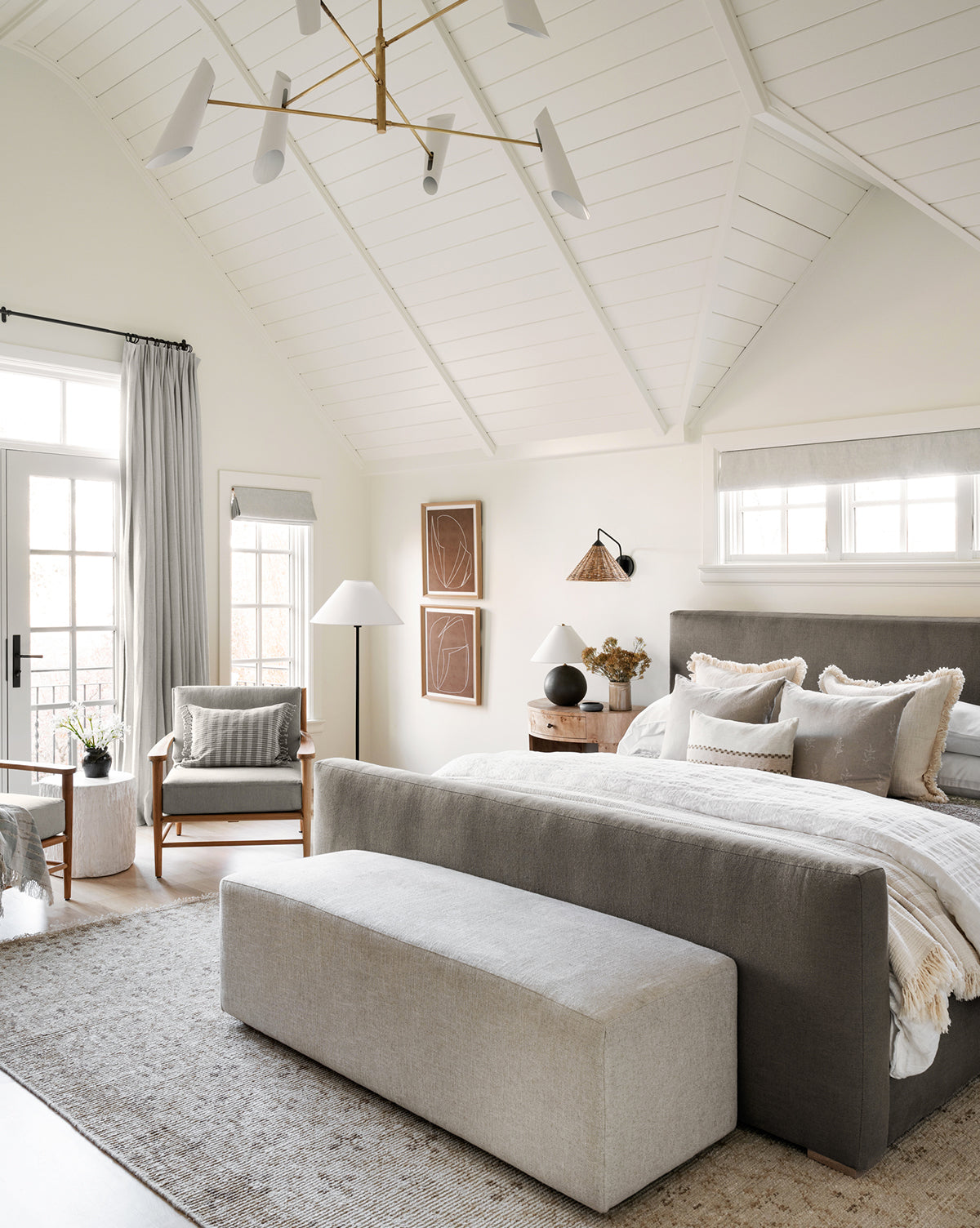 A modern, airy bedroom with vaulted white ceilings, neutral tones, a large bed with gray headboard, matching bench, armchair, wooden nightstand, soft lighting, wall art, and the Mali Hand-Knotted Rug enhancing natural light from windows and glass doors, by Vendixs