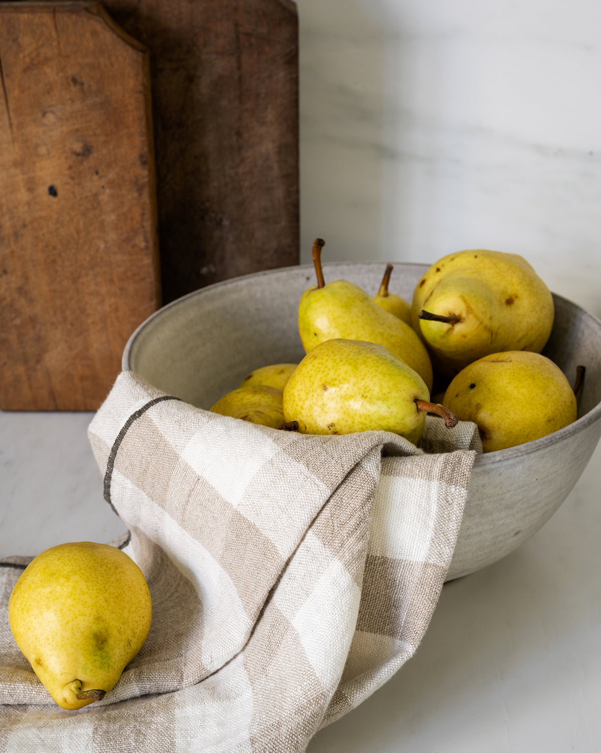 A bowl of yellow pears sits on a countertop, partially covered with the Charlie Check Tea Towel. One pear rests outside the bowl, while wooden boards in the background create a cozy table setting, by Vendixs