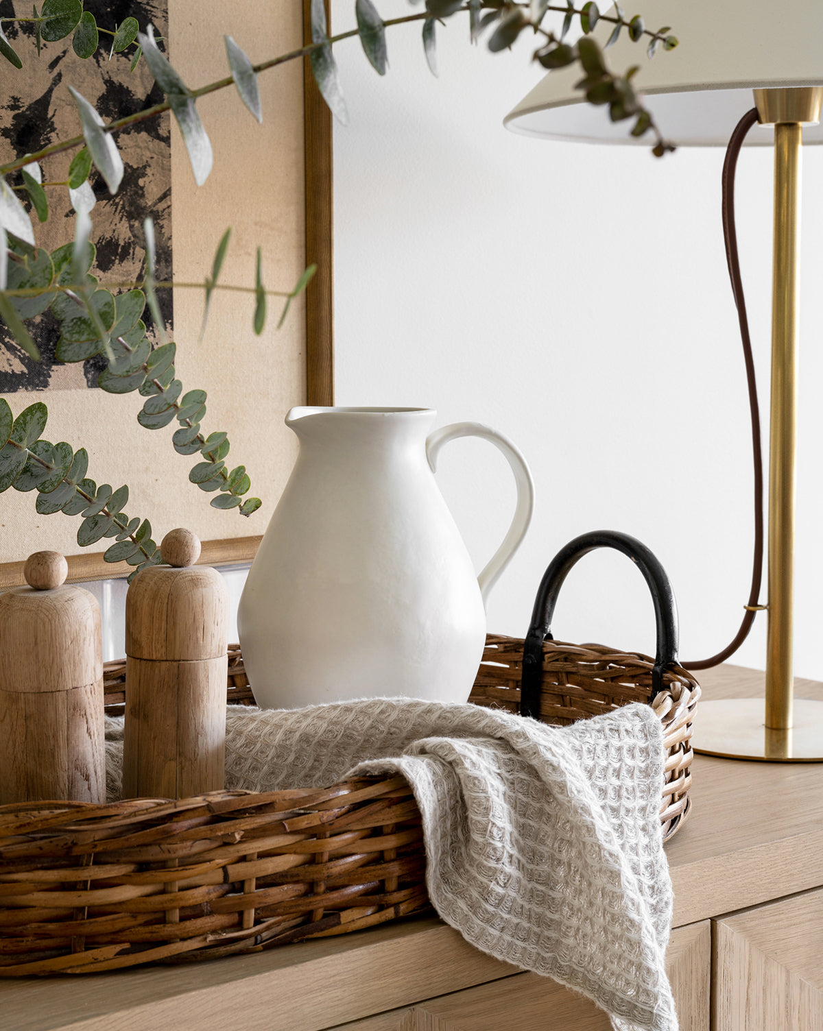 A wicker tray on wood holds a White Ceramic Pitcher, two wooden grinders, and a textured cloth; eucalyptus branches and a brass lamp sit nearby, by Vendixs