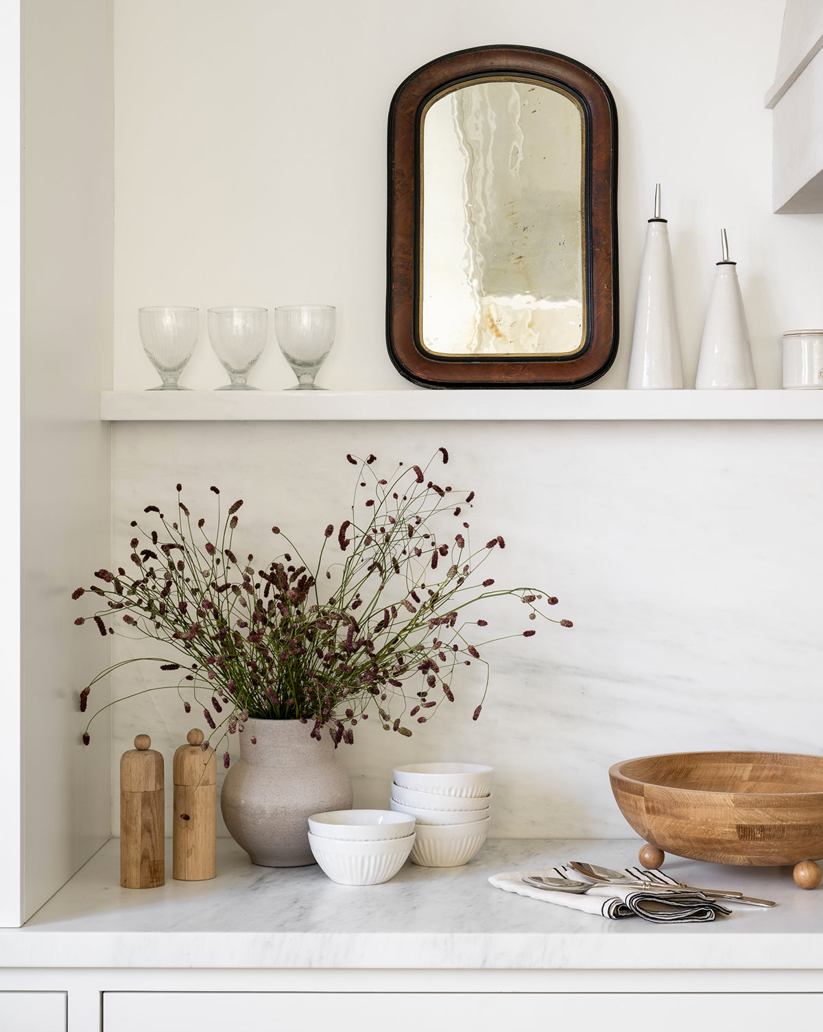A minimalist kitchen shelf features a rustic mirror, fluted glassware, tall ceramic bottles, wildflowers in a vase, wooden bowls, stacked Adele Bowls in white, salt and pepper mills, and arranged cutlery on a marble countertop by Vendixs