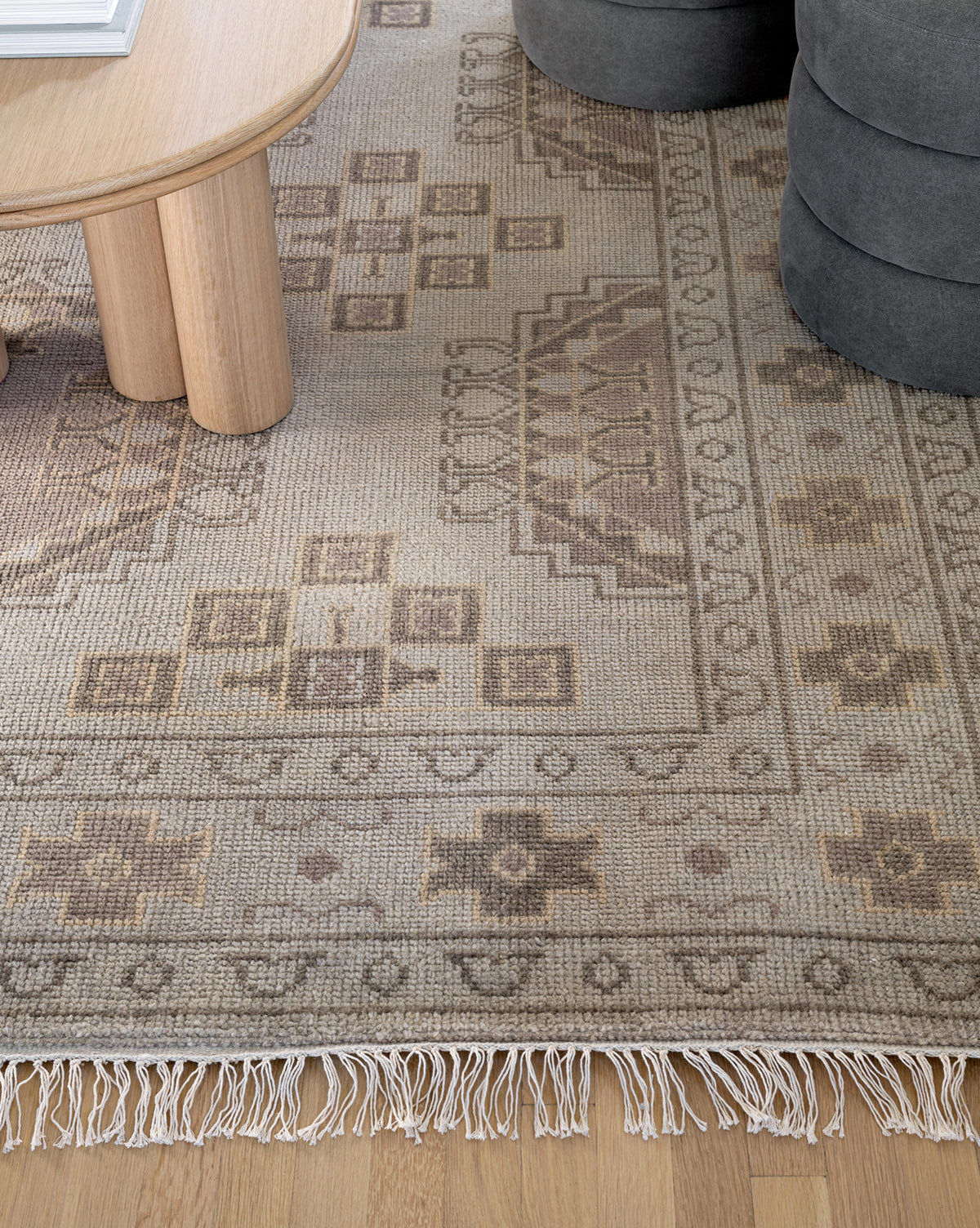A close-up of the Kenna Hand-Knotted Wool Rug, featuring beige and brown geometric patterns with fringe, partially placed under a round wooden table beside a gray ottoman on a light wood floor, by Vendixs