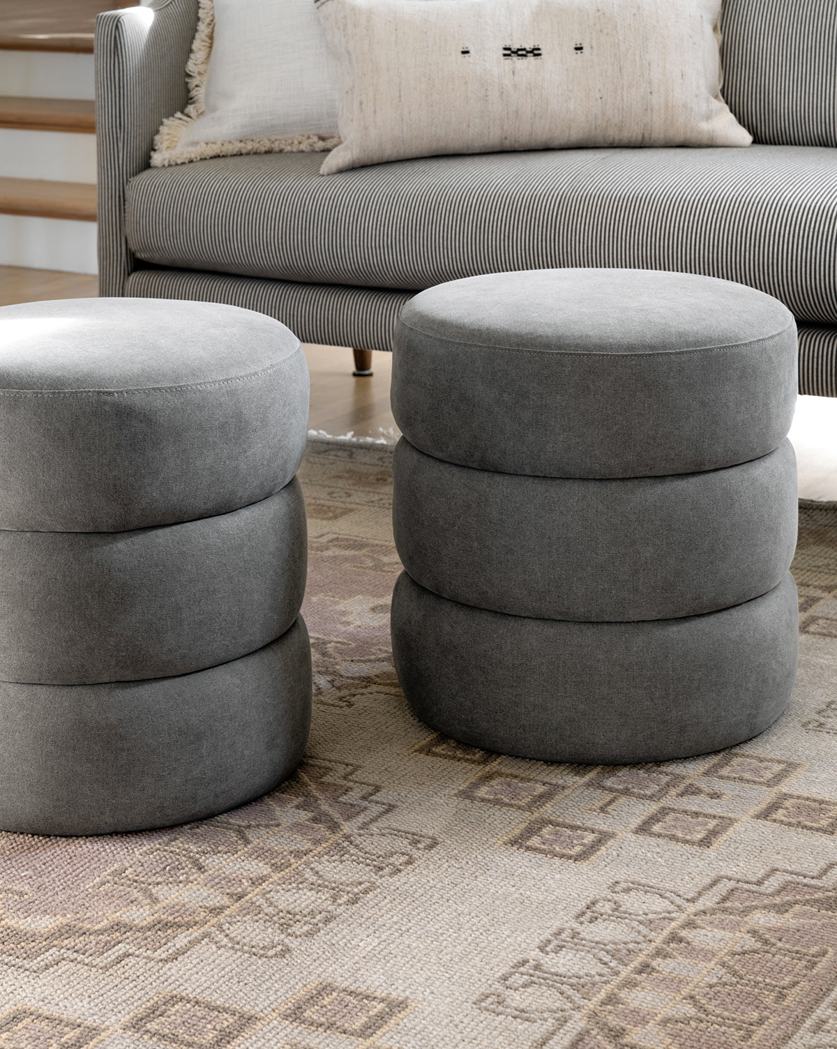 Two round gray Channel Ottomans with stacked cushion-like sections sit on a patterned rug in front of a striped sofa with neutral pillows, adding midcentury flair to the space, by Vendixs