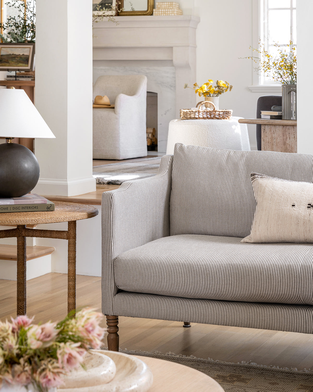 A cozy living room showcases the Makerspalm Gemma Settee 65 in striped fabric with a neutral pillow, paired with a side table, lamp, books, and flowers—complementing the classic fireplace and armchair in the background, finished with touches from Vendixs