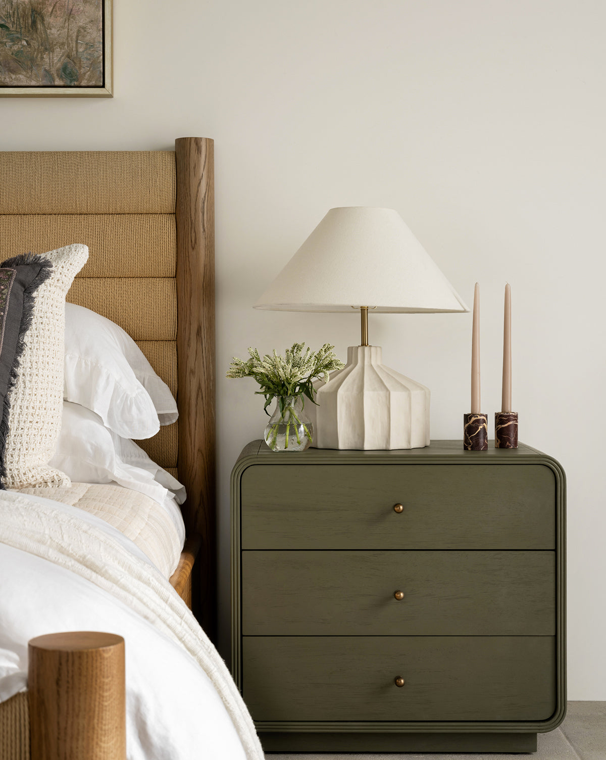 A neatly made bed sits beside a green three-drawer nightstand, which is topped with a Veneto Table Lamp, two tall candles in holders, and a small vase of flowers, all set against a plain white wall by Vendixs