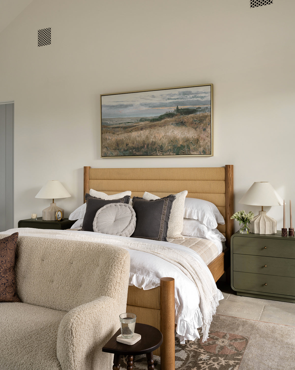 A cozy bedroom with a tan upholstered bed, layered pillows and blankets, two green nightstands with lamps, a beige textured sofa, a landscape painting above the bed, and the Suzani Hand-Knotted Wool Rug adding warmth to the space, by Vendixs