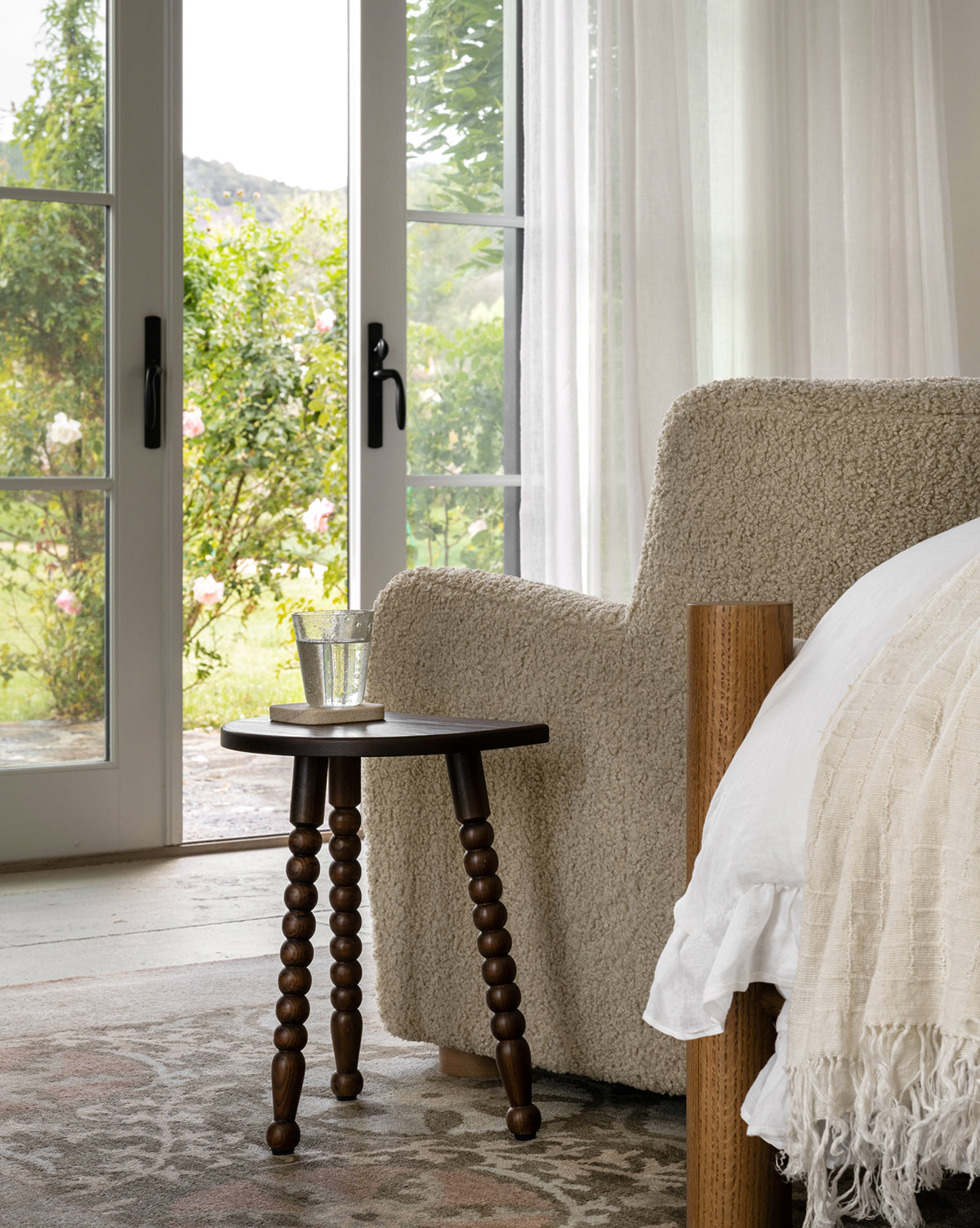 A cozy beige armchair and a white bed sit by glass doors, complemented by the Alice Wood Stool in ash. Sheer curtains and natural light create a serene, inviting atmosphere with views of the lush garden by Vendixs