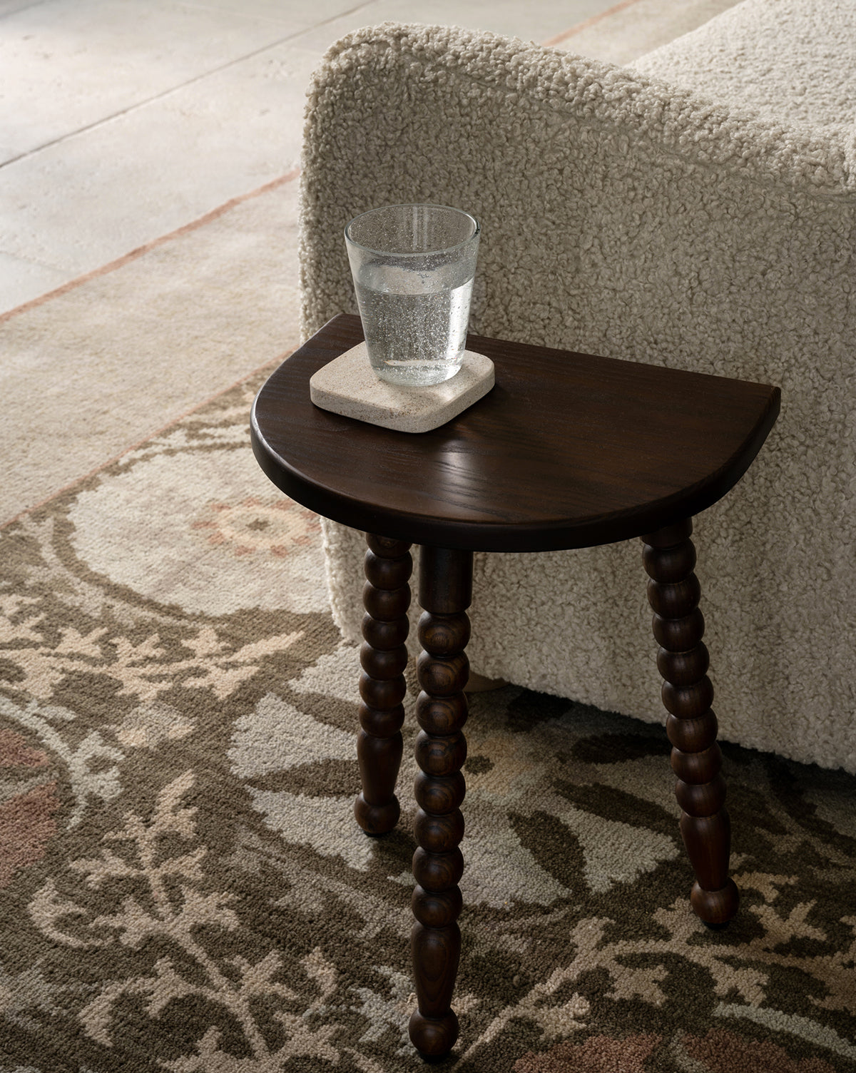 A small dark wood side table with carved legs holds a glass of water on a stone coaster beside a textured cream sofa, all set atop the Suzani Hand-Knotted Wool Rug, by Vendixs