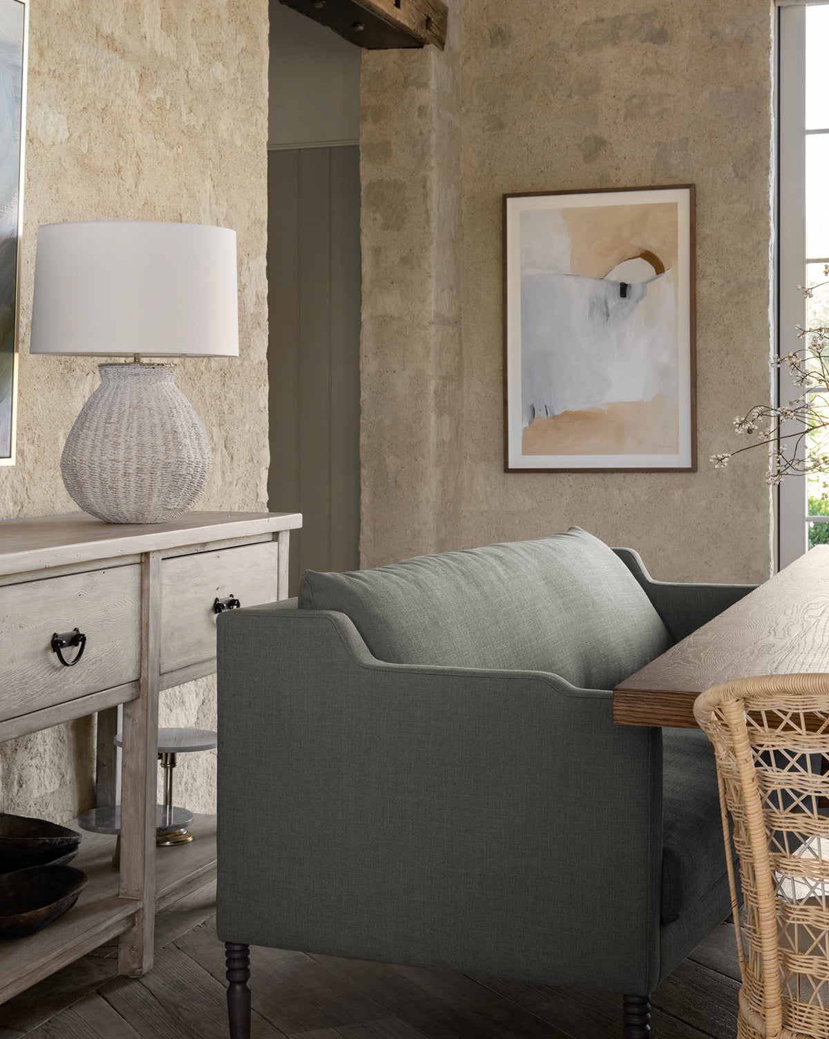 A cozy living room features the Makerspalm Gemma Settee 65 in high-quality fabric, paired with a light wooden sideboard, textured white lamp, abstract wall art, and a rustic stone wall for a warm, inviting atmosphere—finished with thoughtful touches inspired by Vendixs