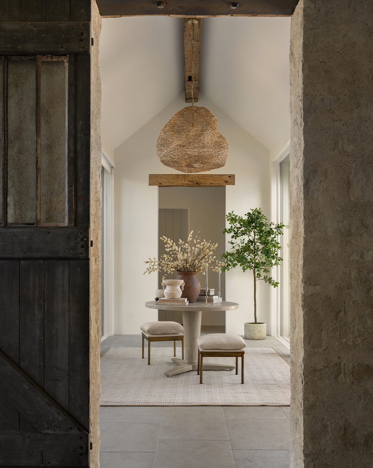 A rustic dining area with a round table, two stools, a large vase with branches, a potted tree, and organic lighting from the Andorra Wicker Pendant. The room features stone walls, wooden beams, and neutral tones, by Vendixs