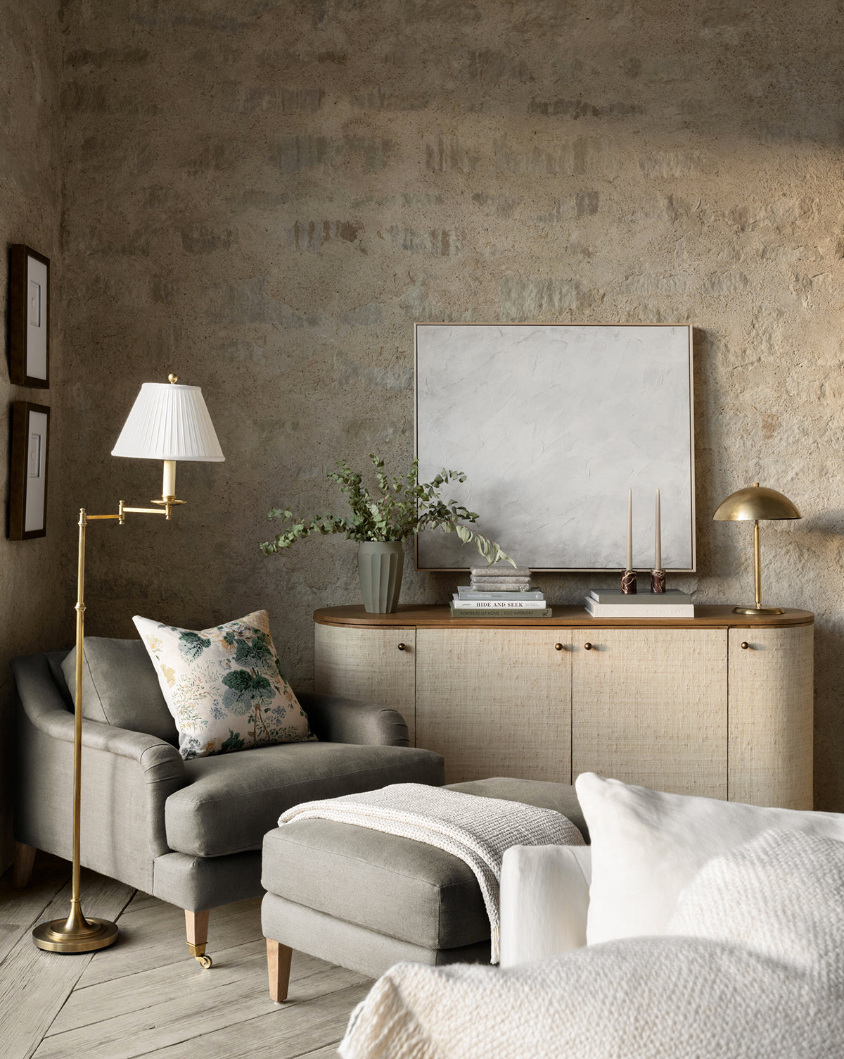 A cozy living room corner features a gray armchair, floral pillow, Lucille Ottoman, and cream throw. Nearby, a sideboard displays books, a vase with greenery, candles, and a gold lamp under an abstract painting on a stone-textured wall by McGee & Co.