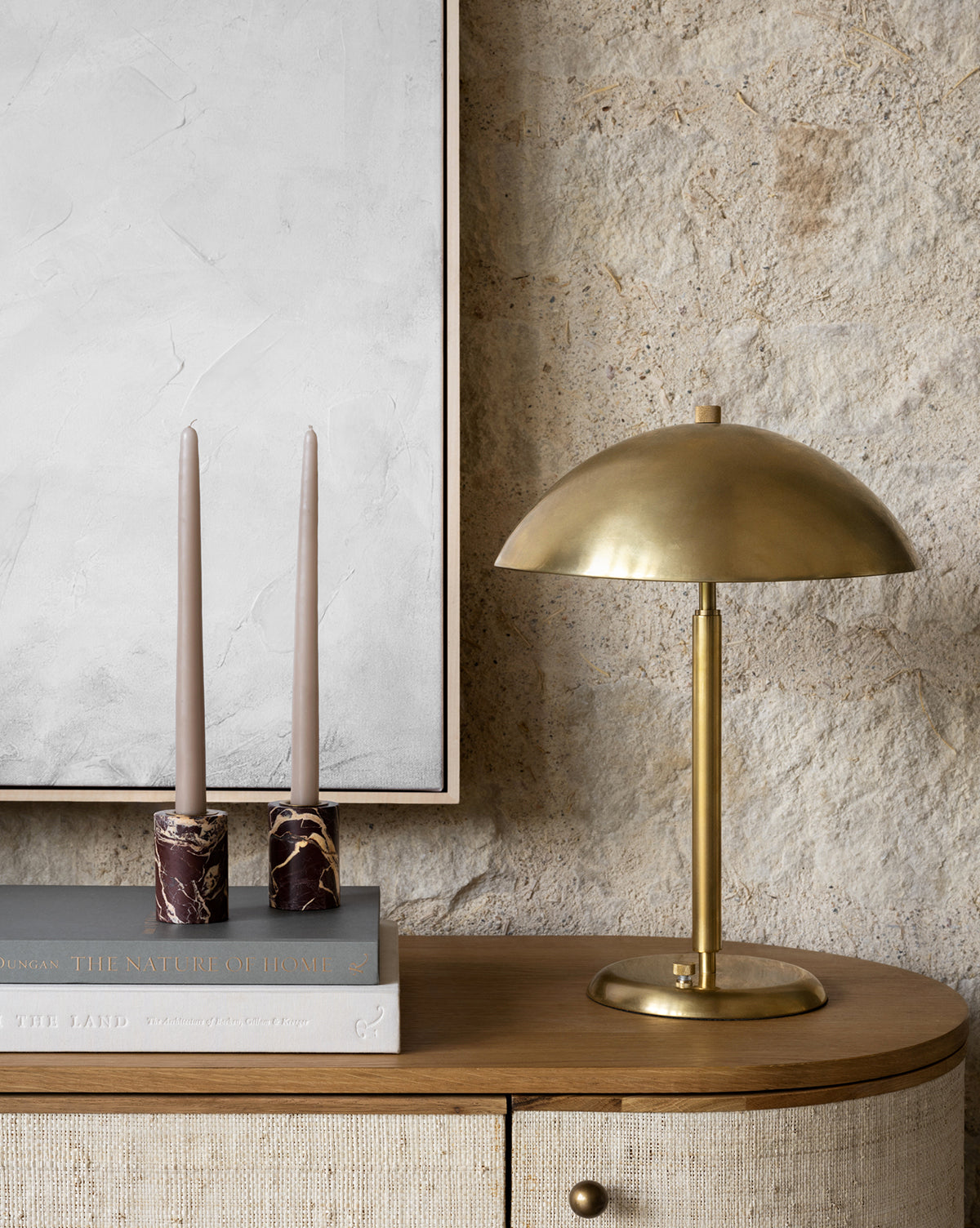 The Aldorno Desk Table Lamp sits on a wooden cabinet beside two tall beige candles in marble holders and books, under white textured wall art against a stone wall—an ideal choice for chic living room lighting, by Vendixs