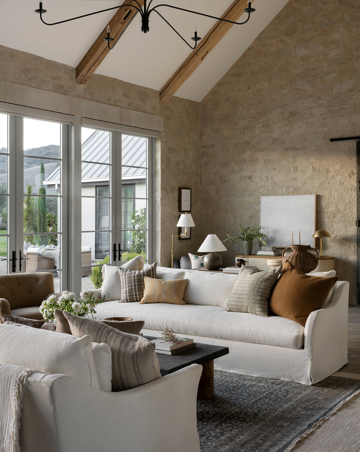 A modern living room features high vaulted ceilings, beige stone walls, large windows overlooking greenery, white sofas with neutral and brown pillows, a black coffee table, and the Marta Hand-Knotted Wool Rug accenting the space, by Vendixs