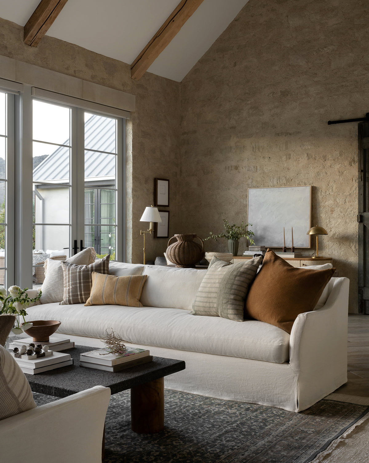 A cozy living room features the Rowe Fine Furniture Wilhelmina Classic Slipcover Sofa (85-110) with neutral and brown pillows, a dark coffee table, greenery, large windows, stone walls, wood beams, minimalist decor, and abundant natural light, blended seamlessly with pieces from Vendixs