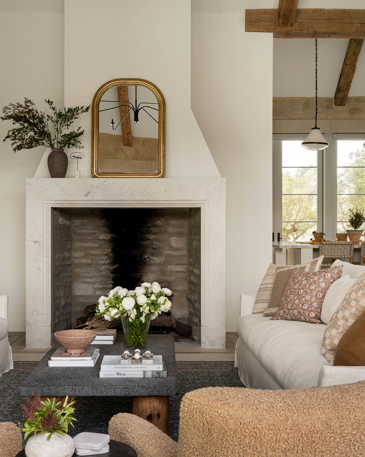 A cozy living room in neutral tones features a stone fireplace with the Giada Wall Mirror above it, a dark coffee table topped with flowers and books, two sofas with patterned pillows, and exposed wooden ceiling beams for an antique touch, by Vendixs