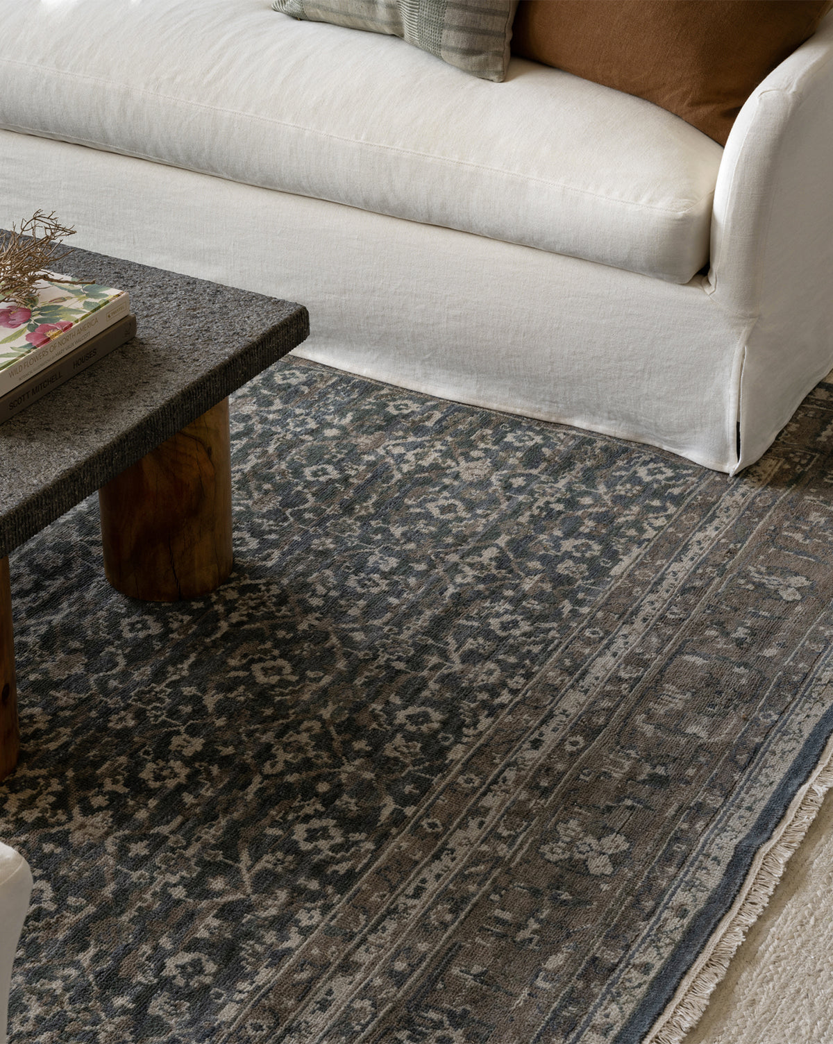 A close-up of a living room featuring the Marta Hand-Knotted Wool Rug, a granite coffee table with a book, and a white sofa adorned with beige and brown throw pillows, by Vendixs
