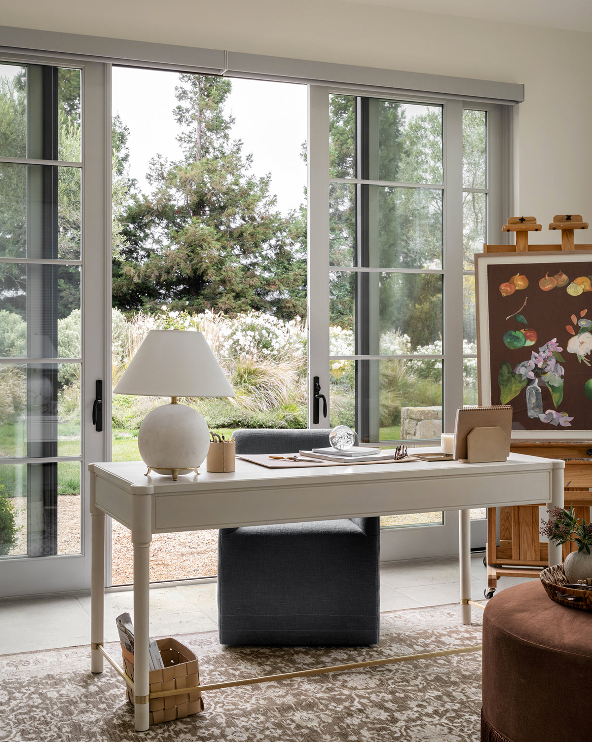 A bright, elegant workspace with a white desk and chair features the Price Table Lamp, its alabaster base and linen shade beside large glass doors. An easel displays a floral painting as sunlight fills the room overlooking a garden, by Vendixs