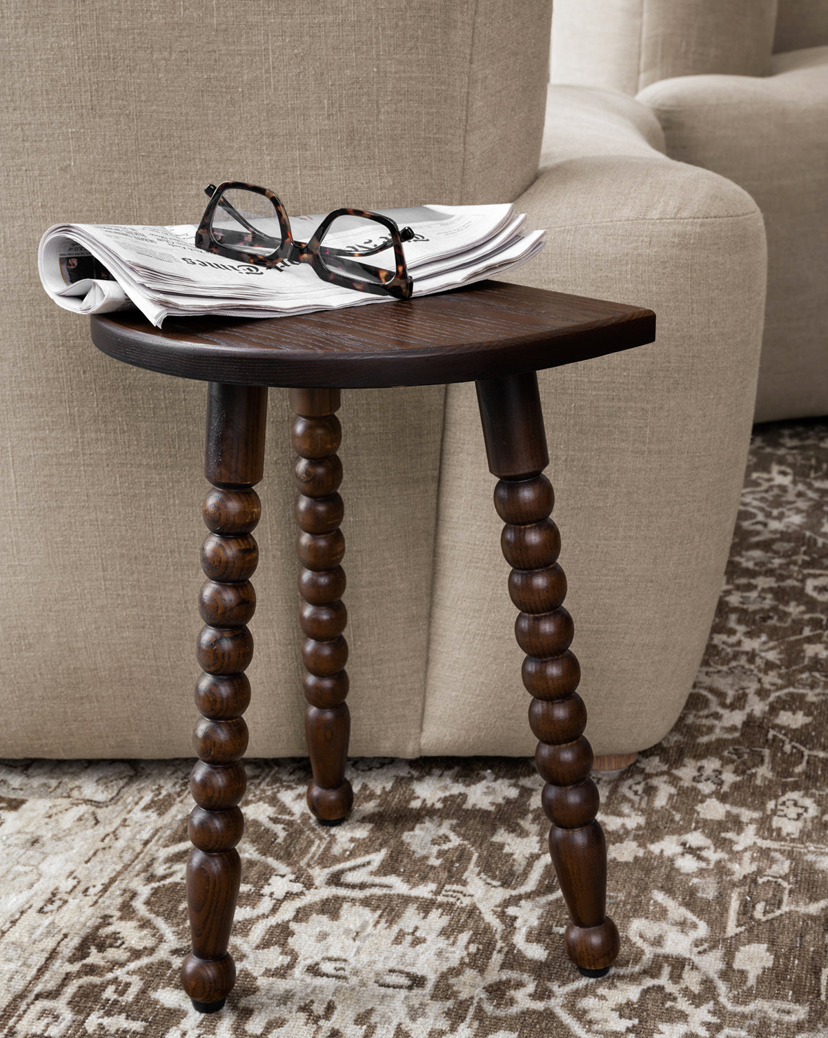 The Alice Wood Stool with three beaded ash wood legs holds a folded newspaper and eyeglasses beside a beige upholstered sofa on a patterned rug, by Vendixs