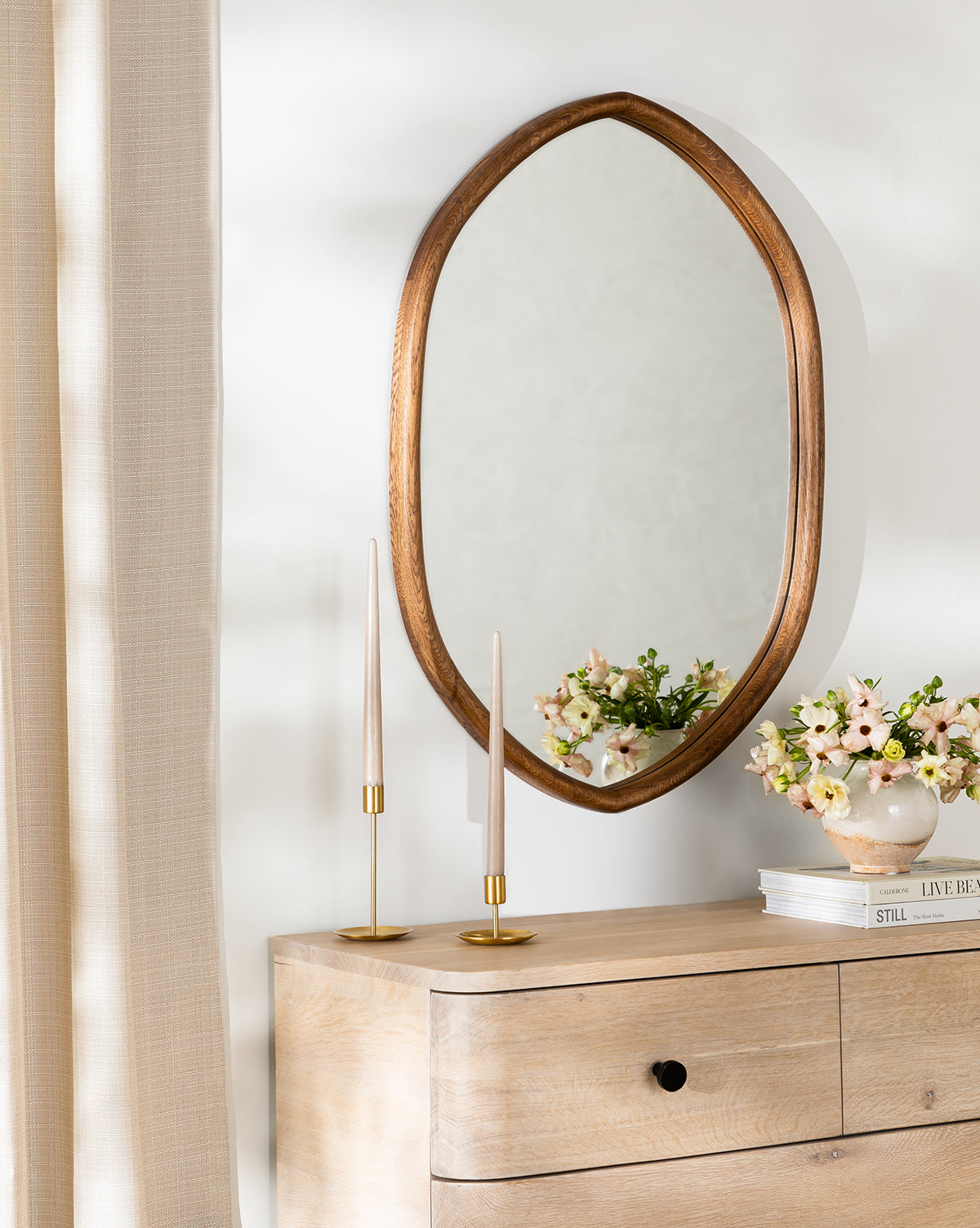 The Oslo Mirror with an oak frame hangs above a wooden dresser decorated with gold candlesticks, a pink flower vase, and a bowl, beside beige curtains and against a white wall, by Vendixs