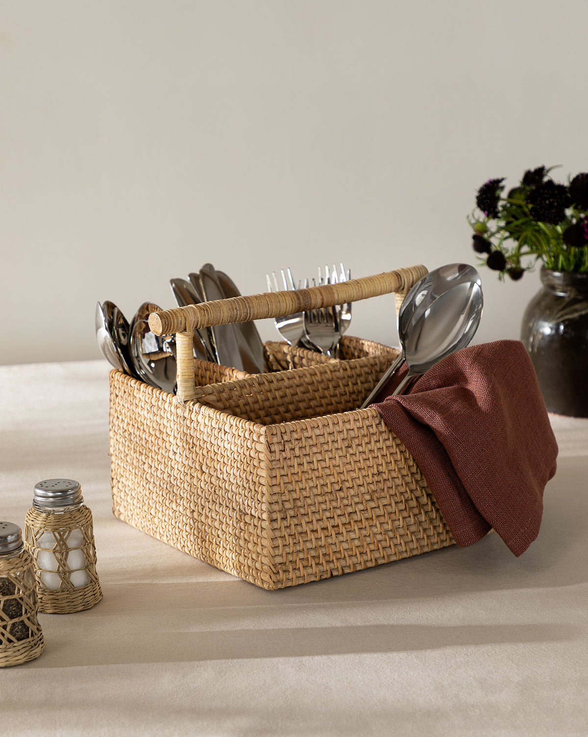 The Rattan Utensil Caddy with a handle stores silverware and a brown napkin. Salt and pepper shakers are nearby, and a dark vase with flowers sits in the background on a beige tablecloth, by Vendixs