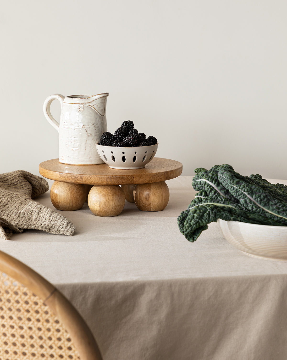 A White Stoneware Pitcher sits on a round wooden stand atop a beige tablecloth, next to a bowl of blackberries, leafy greens, and a nearby wicker chair, by Vendixs
