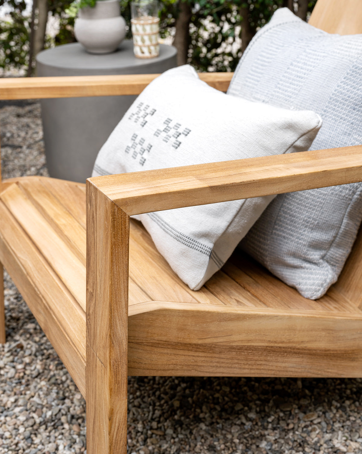 A close-up of the Triby Teak Outdoor Lounge Chair with two textured light gray pillows sits on a gravel patio beside a round gray side table holding a vase and glass, with green foliage in the background, by Vendixs