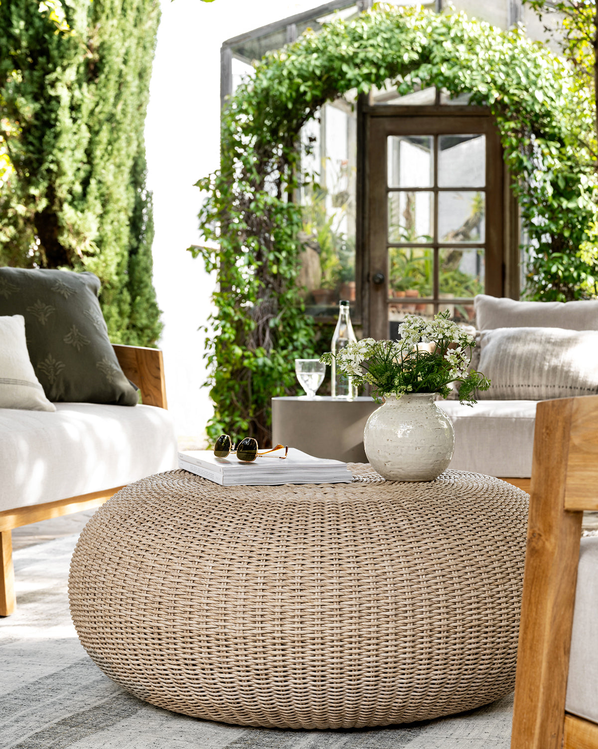 A cozy patio features beige cushioned chairs and the Rimma Outdoor Coffee Table, crafted from all-weather wicker, topped with a white vase of flowers and a book. Lush green ivy arches and a glass door frame the sunlit garden beyond, by Vendixs