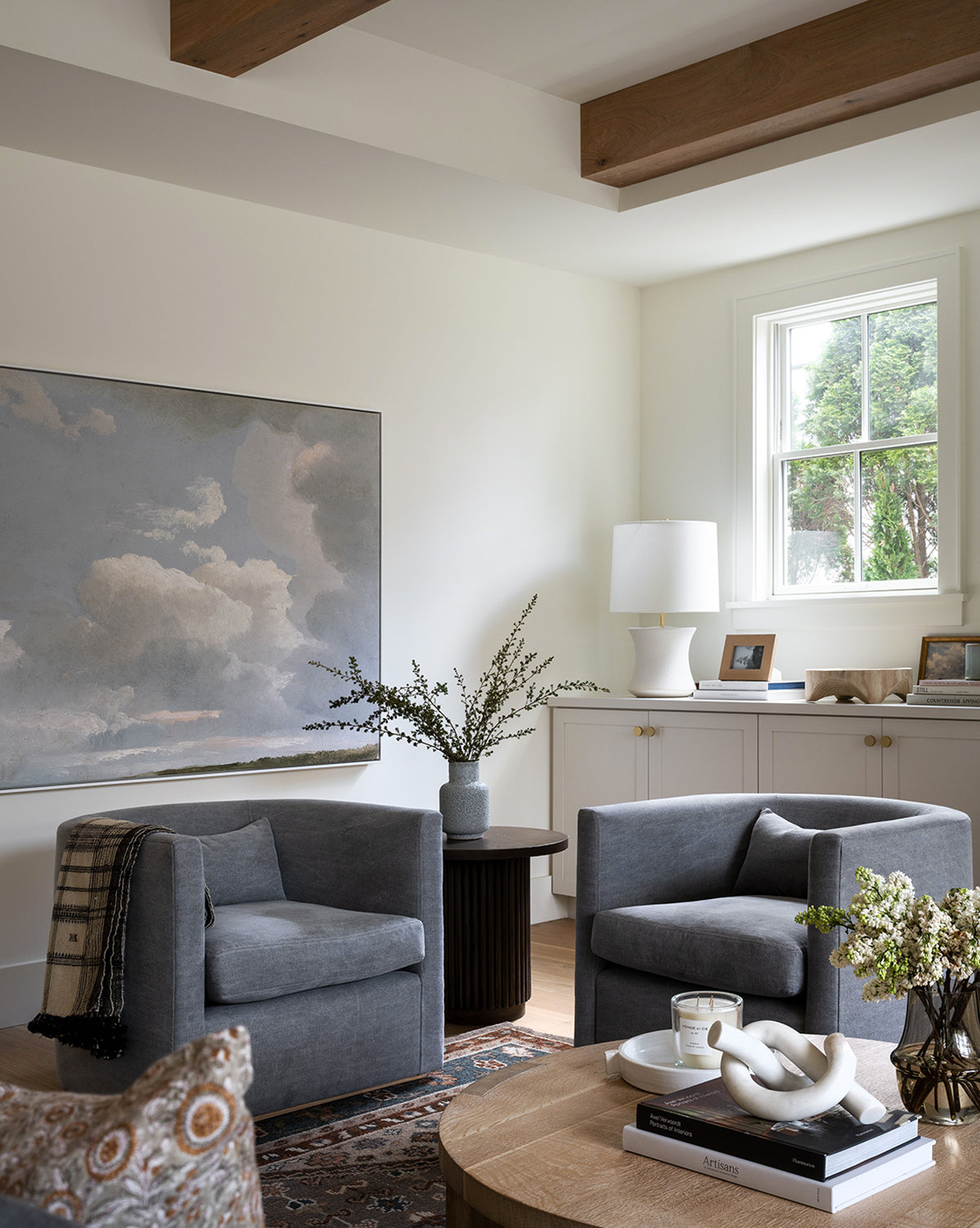 A modern living room with two paired Reese accent chairs featuring down-wrapped cushions, a round handcrafted alder wood coffee table, a large cloudy sky painting, a light-filled window, books, plants, and a decorative lamp by McGee & Co.