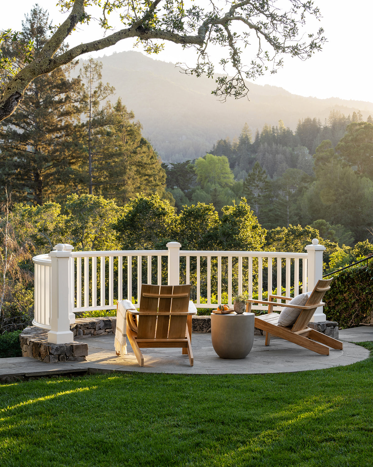 Two Triby Teak Outdoor Lounge Chairs sit on a patio facing a white railing, separated by a small table. Trees and distant mountains are softly illuminated by sunlight beyond this all-weather arrangement, by Vendixs