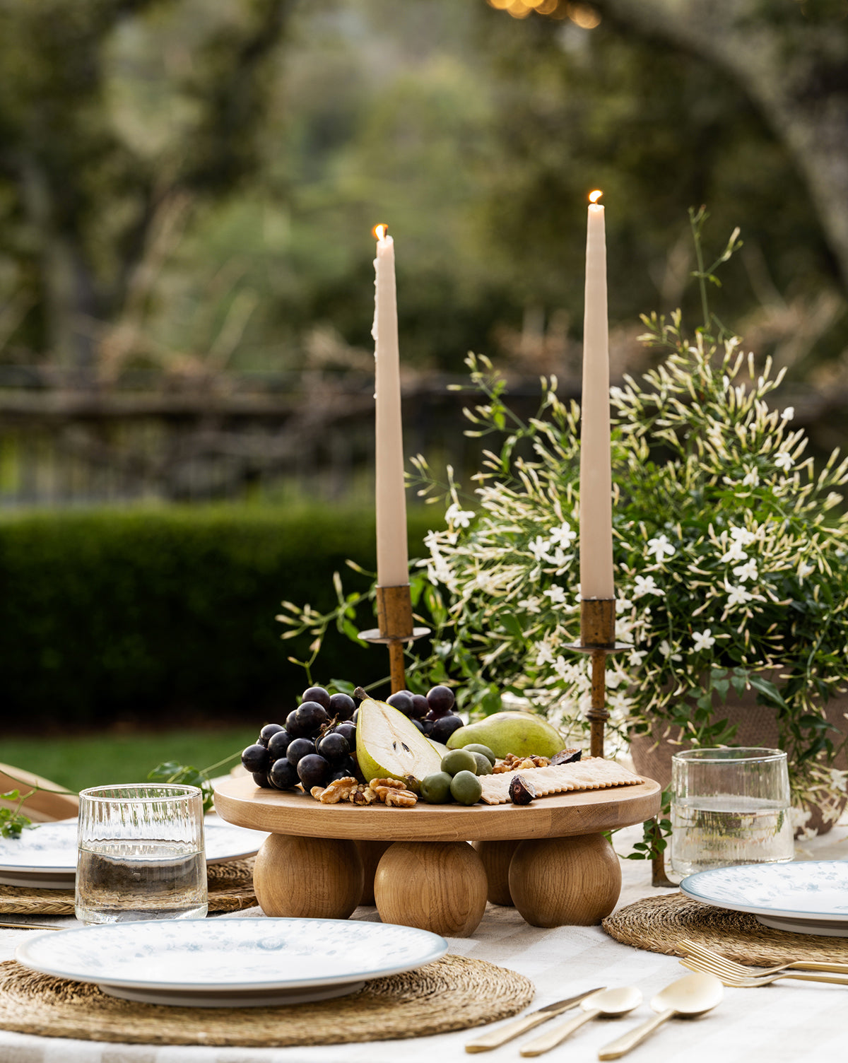 A rustic outdoor table is set with two lit candles, a Bronx Oak Pedestal displaying assorted fruits, cheese, and nuts, two place settings with glassware, and greenery in the background by Vendixs