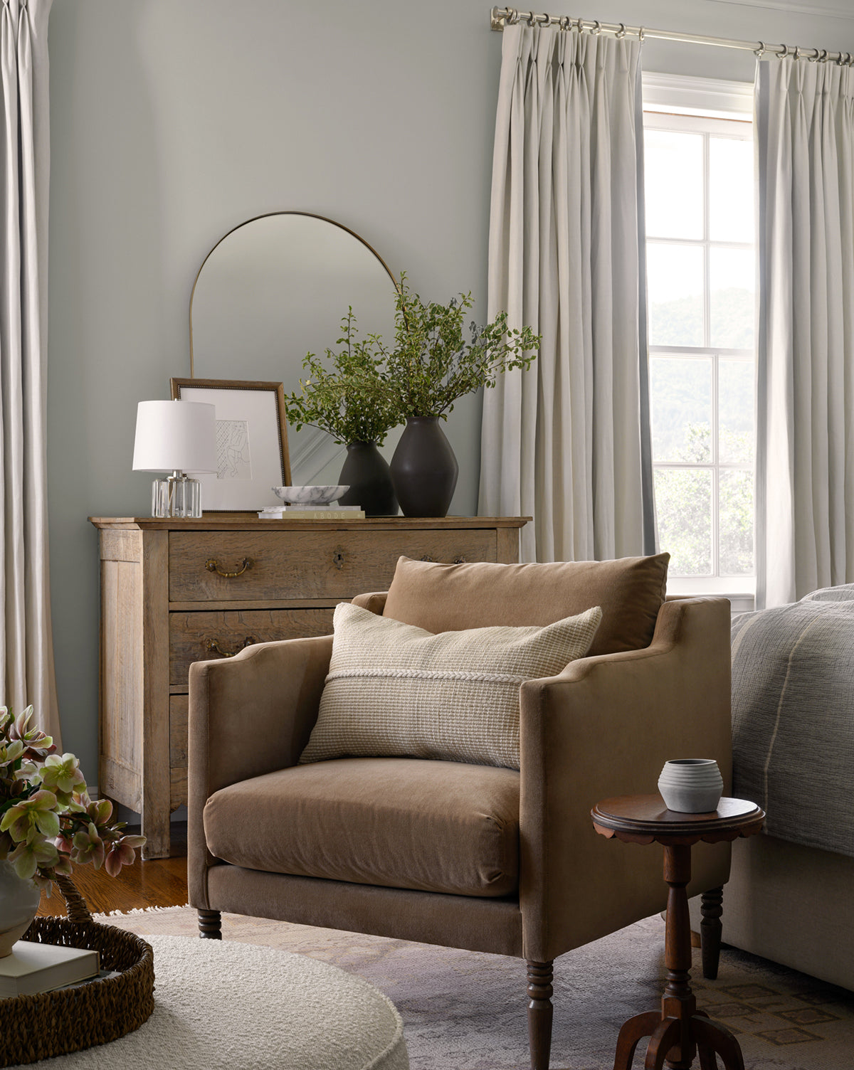 A cozy bedroom corner with the Makerspalm Gemma Lounge Chair, soft pillows, a round side table, a rustic wooden dresser adorned with vases and decor, light curtains, and a large window filling the space with natural light—McGee & Co.