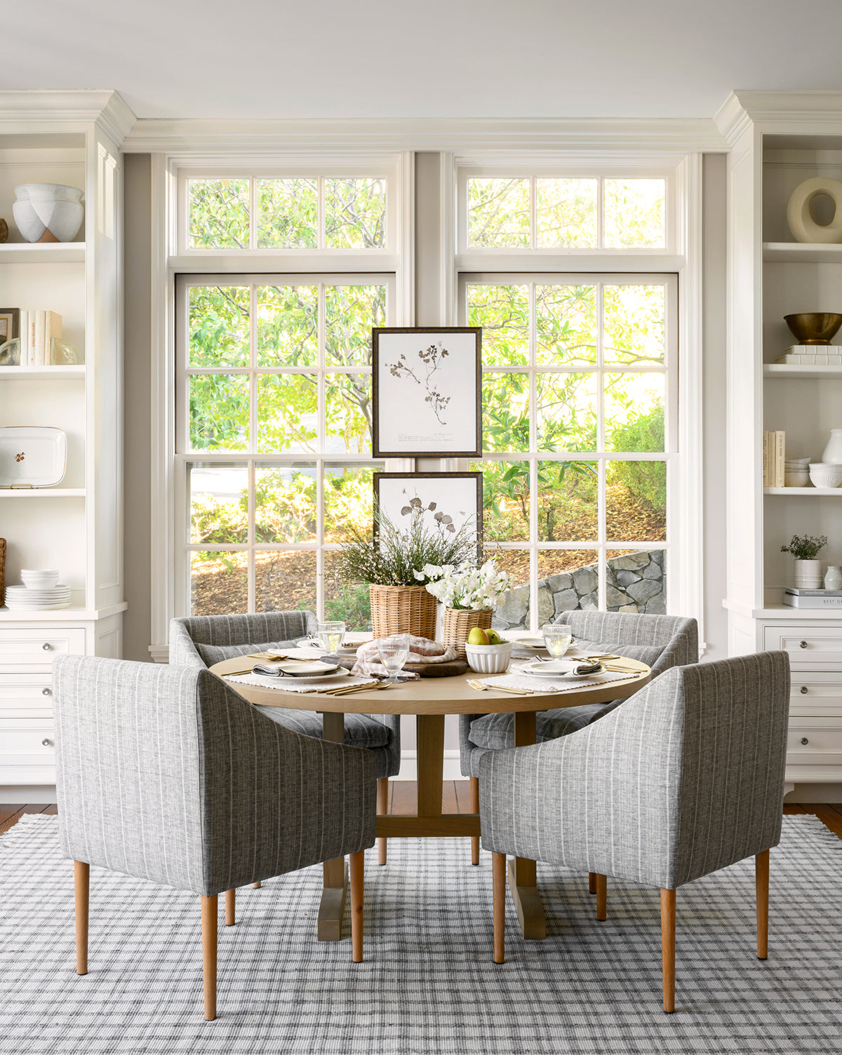 A bright dining area with large windows showcases a Giselle Round Table in white oak set for four, gray upholstered chairs, a white floral centerpiece, and decorated shelves, with greenery visible through the windows by McGee & Co.