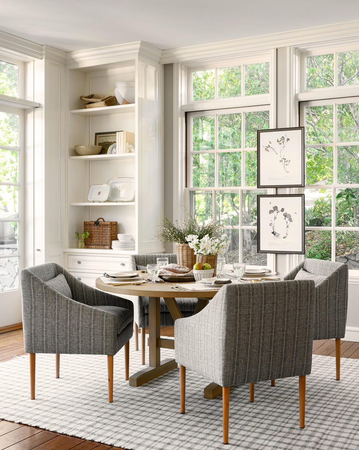 A bright dining room with large windows, a round wooden table for four, gray chairs, a white rug, built-in shelves with decor, and Subdued Botanicals I in a light walnut frame displayed on the wall.