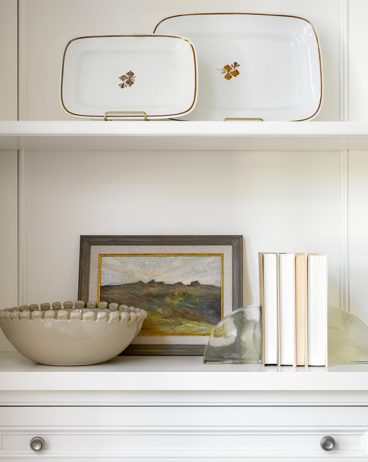 A white shelf holds two decorative plates, the Malibu Bowl, a framed landscape painting, books in a row, and two glass bookends. The all-white background and furniture give a clean look ideal for showcasing unique kitchen décor by Vendixs