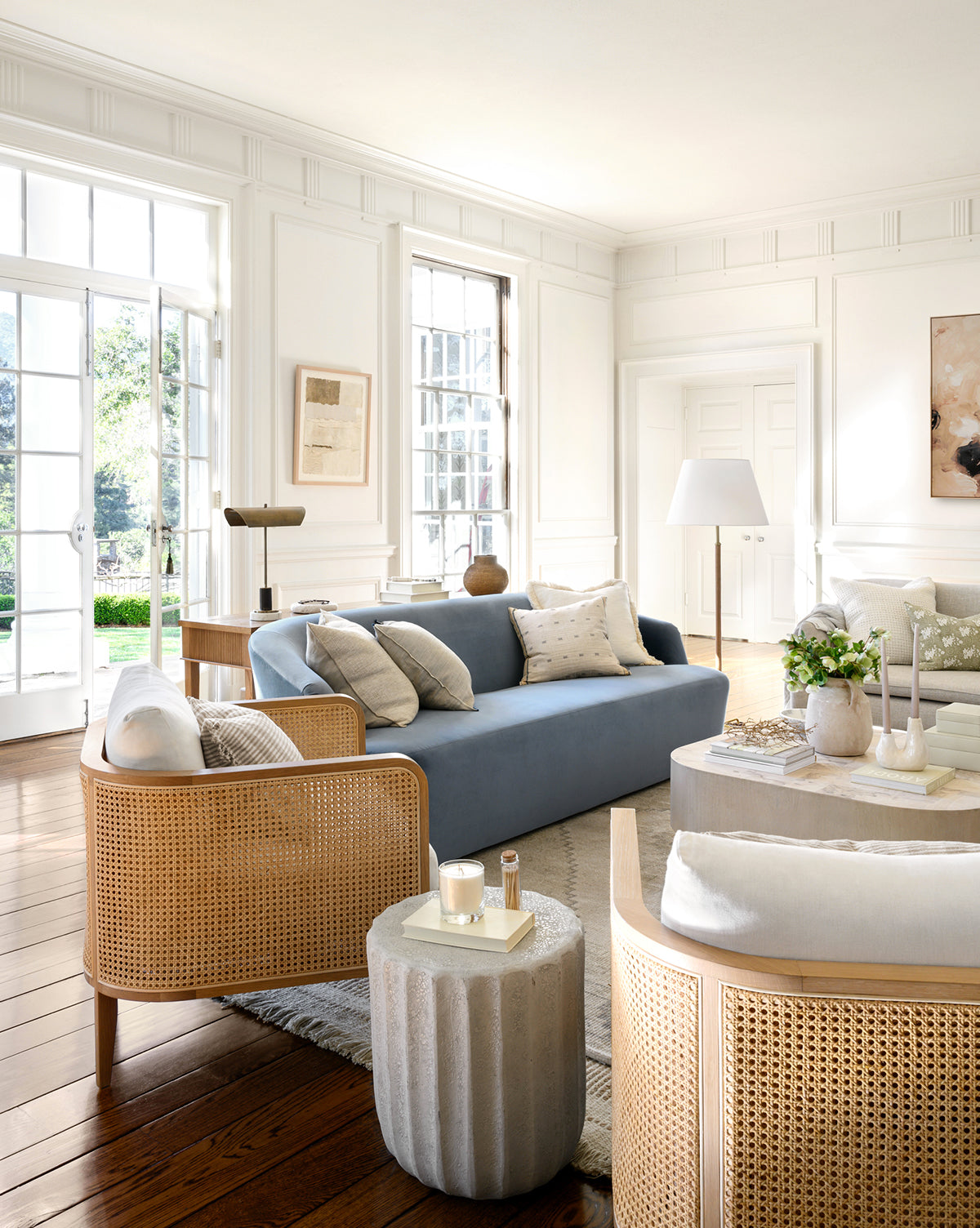 Bright living room with large windows, a Makerspalm Byron Sofa 96 in blue, rattan armchairs, a light rug, and modern decor. Sunlight highlights wood floors and neutral accents, with a lush garden view outside—finished with touches from Vendixs